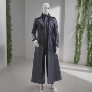 May include: A black leather trench coat with a matching zip-up jacket and pants. The coat features a long, flowing design with a button-up front and a belt detail on the sleeves. The mannequin is standing in a well-lit room with green plants.