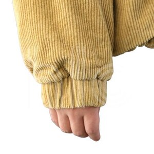 Puede incluir: Primer plano de una manga de chaqueta de pana amarillo mostaza. La manga est&aacute; recogida en la mu&ntilde;eca con un pu&ntilde;o el&aacute;stico. La tela de pana tiene una textura acanalada vertical.