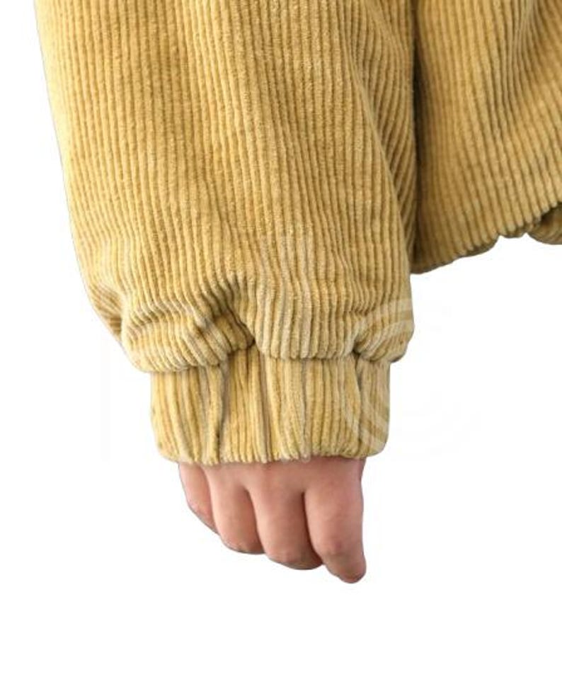Puede incluir: Primer plano de una manga de chaqueta de pana amarillo mostaza. La manga est&aacute; recogida en la mu&ntilde;eca con un pu&ntilde;o el&aacute;stico. La tela de pana tiene una textura acanalada vertical.