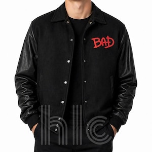 May include: Black varsity jacket with black leather sleeves and a button-up front. The word "BAD" is embroidered in red on the chest. The jacket has a classic collar and ribbed cuffs.