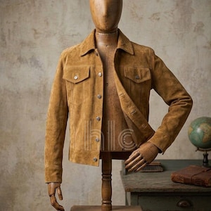 Könnte beinhalten: Hellbraune Wildlederjacke auf einer Holzpuppe. Die Jacke hat einen klassischen Kragen, zwei Brusttaschen und Knopfverschlüsse. Die Puppe steht vor einem Hintergrund im Vintage-Stil, mit einem Globus und einem Buch im Hintergrund.