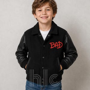 May include: Black varsity jacket with leather sleeves and the word "BAD" embroidered in red. The jacket has a button-up front and a classic collar. The jacket is worn with blue jeans.