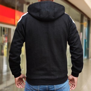 May include: Black hooded sweatshirt with white stripes on the shoulders. The sweatshirt has a drawstring hood and ribbed cuffs and hem. The person is wearing blue jeans.