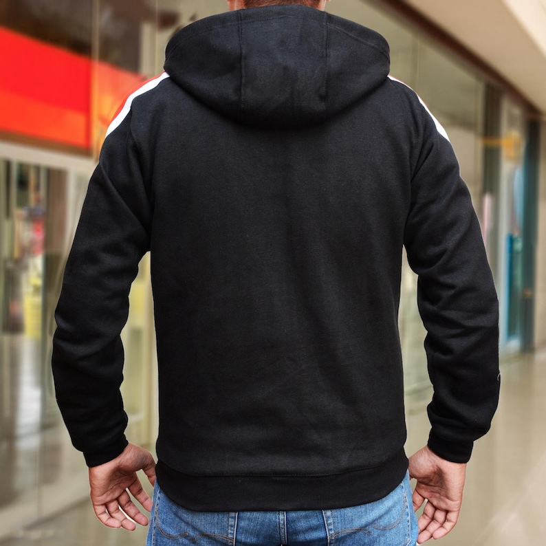 May include: Black hooded sweatshirt with white stripes on the shoulders. The sweatshirt has a drawstring hood and ribbed cuffs and hem. The person is wearing blue jeans.