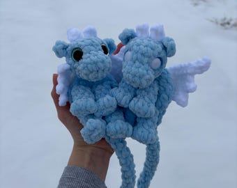 Puede incluir: Dos peluches de dragón azul tejidos a crochet con alas blancas y ojos saltones. Los dragones están sentados en una mano.