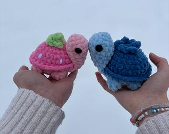 May include: Two crocheted turtle plushies, one pink with a green leaf on its shell and one blue, are held in the hands of a person. The turtles are facing each other.