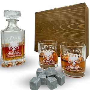 Könnte beinhalten: Ein Holzkasten-Set mit einer Glaskaraffe und zwei Gläsern. Die Karaffe und die Gläser sind mit dem Text "Leano Albania All Original Parts" und einem stilisierten Adler graviert. Das Set enthält außerdem sechs graue Stein-Eiswürfel.