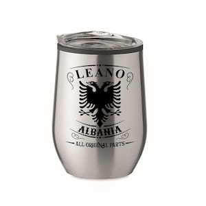 Könnte beinhalten: Ein isolierter Edelstahlbecher mit schwarzem Deckel. Der Becher hat eine schwarze Grafik eines doppelköpfigen Adlers, das Wort "LEANO" darüber und "ALBANIA" darunter, mit "ALL ORIGINAL PARTS" unten.
