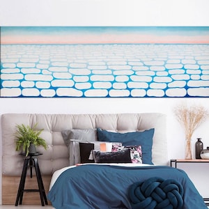 May include: A bedroom interior with a large abstract painting of a blue and white sky above a white brick pattern. The painting is hanging above a bed with a blue duvet and a blue pillow. There is a small black side table next to the bed with a potted plant.