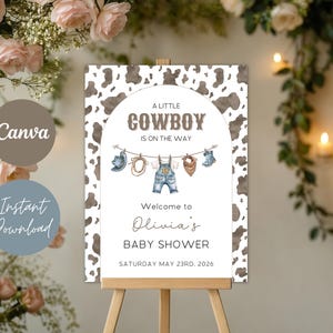 May include: A baby shower sign with a cowboy theme. The sign features the text "A Little Cowboy is on the Way" and "Welcome to Olivia's Baby Shower". The background has a cowhide pattern. The sign is displayed on a wooden easel.