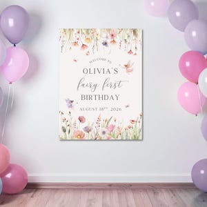 May include: A sign with floral illustrations and the text "Welcome to Olivia's fairy first birthday August 18th, 2026". The sign is surrounded by pink, purple, and white balloons, creating a festive atmosphere.