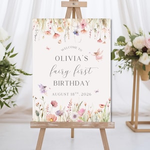 May include: A sign on a wooden easel with the text "Welcome to Olivia's fairy first birthday August 18th, 2026". The sign is decorated with watercolor flowers, butterflies, and a fairy illustration. The background is a soft cream color.