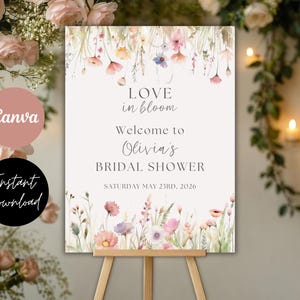 May include: A floral bridal shower sign on a wooden easel. The sign reads "Love in bloom, Welcome to Olivia's Bridal Shower, Saturday May 23rd, 2026." The sign has a watercolor floral design with pink, purple, and yellow flowers.