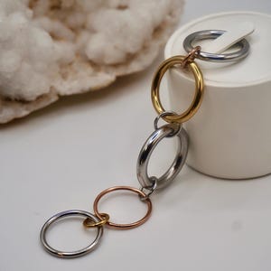 May include: A chain of interlocking metal rings in gold, silver, and rose gold. The rings are connected to each other and are hanging from a white display stand.
