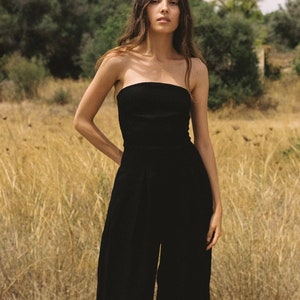 K&ouml;nnte beinhalten: Ein schwarzer, tr&auml;gerloser Jumpsuit mit weiten Beinen. Das Model steht in einem Feld mit hohem Gras.