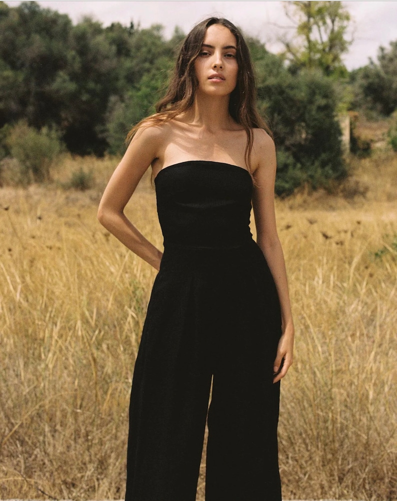 K&ouml;nnte beinhalten: Ein schwarzer, tr&auml;gerloser Jumpsuit mit weiten Beinen. Das Model steht in einem Feld mit hohem Gras.
