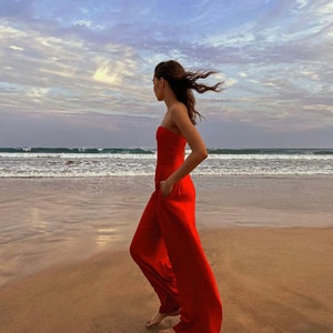 May include: A woman in a red strapless jumpsuit walks on a sandy beach with the ocean in the background. The wind blows her hair.