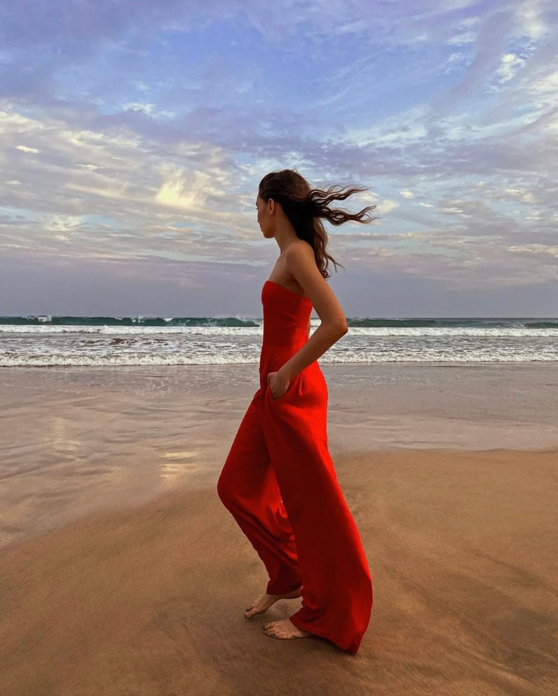 May include: A woman in a red strapless jumpsuit walks on a sandy beach with the ocean in the background. The wind blows her hair.