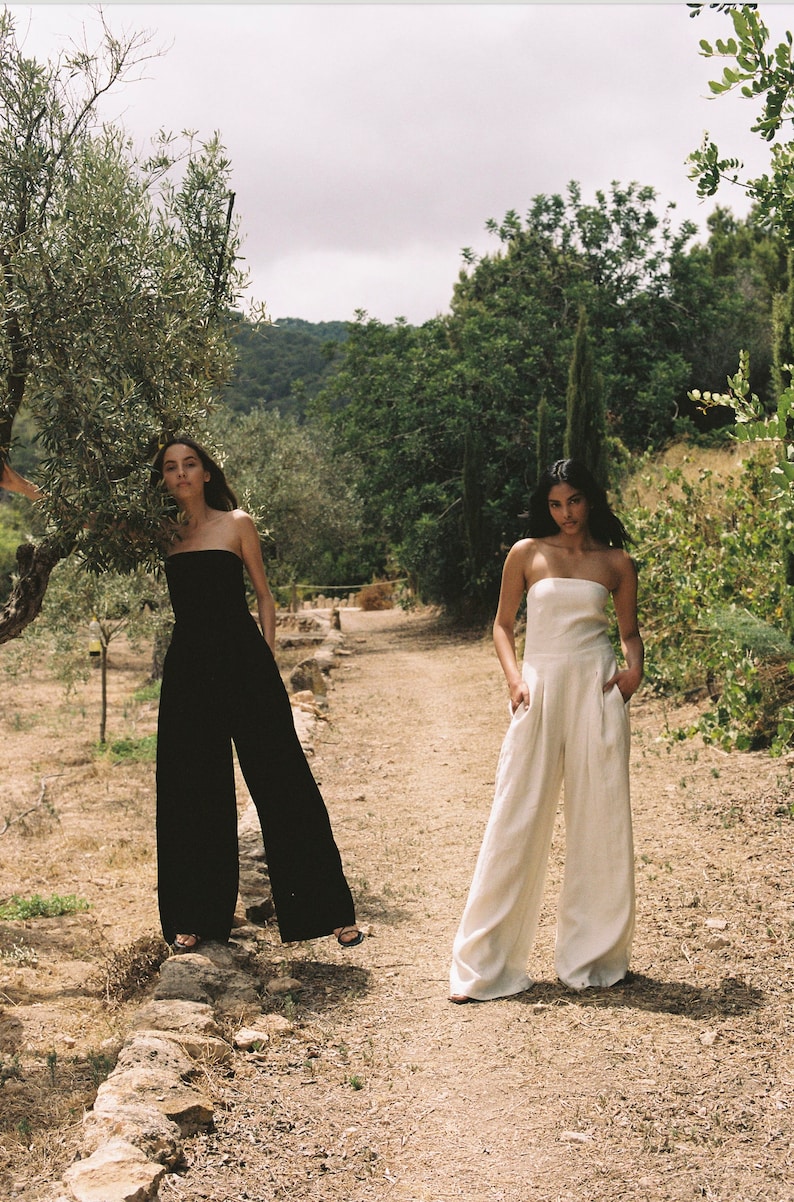 K&ouml;nnte beinhalten: Zwei Frauen in tr&auml;gerlosen Jumpsuits, einer schwarzen und einem wei&szlig;en, stehen auf einem Feldweg in einem Garten.