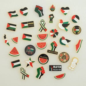 May include: A collection of Palestine-themed pins and badges. The pins feature the Palestinian flag, watermelon slices, doves, and fist symbols. Some pins include the text "FREE PALESTINE" and "PRAY FOR PALESTINE".