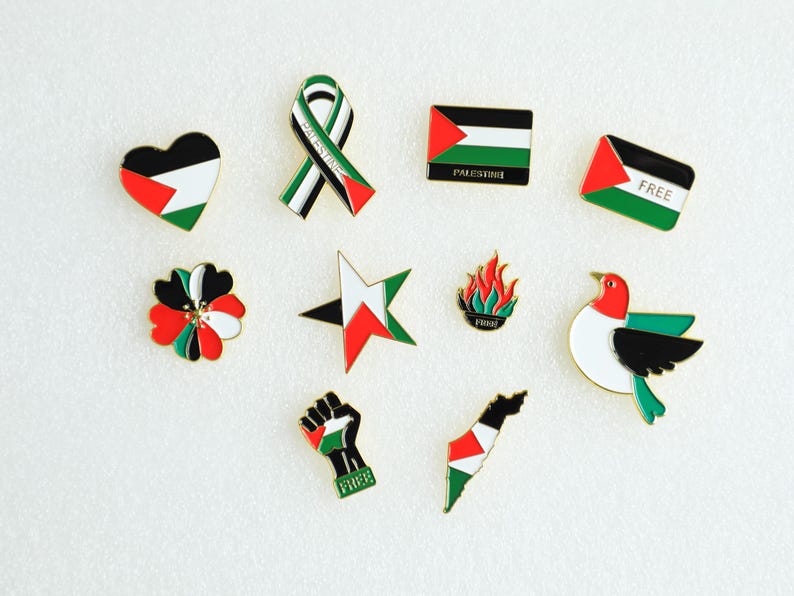 May include: A collection of enamel pins featuring the colors of the Palestinian flag. Designs include a heart, ribbon, flag, star, flower, flame, dove, fist, and a map of Palestine. Some pins have the word "FREE".
