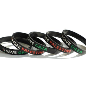 以下が含まれることがあります： 「FREE PALESTINE」と「SAVE GAZA」の文字が白、赤、緑でプリントされた、黒いラバーリストバンドが5つ。白い面に並べられています。