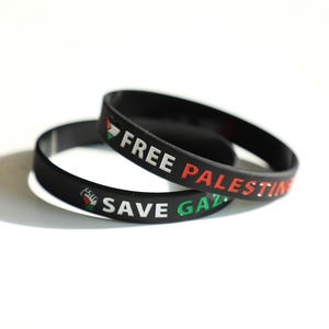 Könnte beinhalten: Zwei schwarze Armbänder aus Gummi mit weißem, rotem und grünem Text. Auf einem Armband steht "FREE PALESTINE", auf dem anderen "SAVE GAZA". Die Armbänder sind übereinander gestapelt.