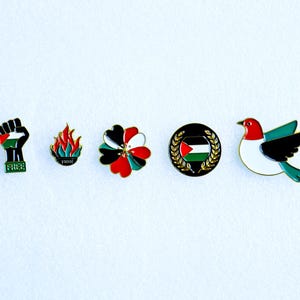 May include: A collection of five enamel pins featuring Palestinian symbols. Designs include a raised fist with the Palestinian flag, flames, a flower, a flag within a wreath, and a bird. The word "FREE" is on the fist pin.