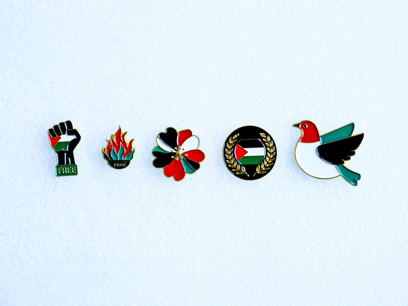 May include: A collection of five enamel pins featuring Palestinian symbols. Designs include a raised fist with the Palestinian flag, flames, a flower, a flag within a wreath, and a bird. The word "FREE" is on the fist pin.