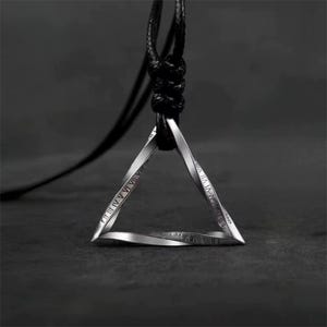 Puede incluir: Un collar con un colgante de triángulo plateado con marcas de números romanos en cada lado. El collar está en un cordón negro.