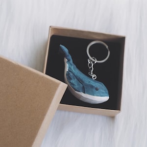 Puede incluir: Un llavero de madera de ballena azul y blanco en una caja marrón. La ballena está tallada con una superficie texturizada y tiene un llavero plateado.