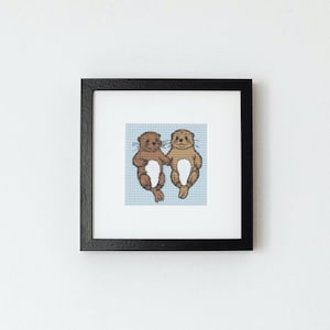 May include: A framed cross-stitch picture showing two cartoon otters holding hands. The otters are brown and tan, set against a light blue checkered background. The artwork is in a black square frame and mounted on a white wall.