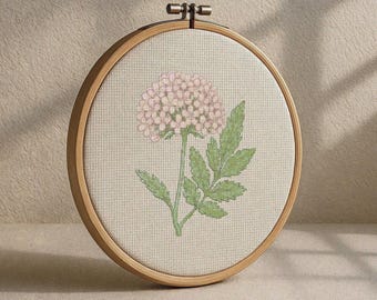 May include: A cross-stitch embroidery in a wooden hoop. The design shows a pink flower with green leaves. The fabric is a light beige colour. The wooden hoop is a light brown colour.