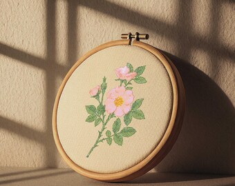 May include: An embroidery hoop with a floral design. The design features pink roses with green leaves and a yellow center, stitched on beige fabric. The fabric is held within a round wooden hoop. The hoop is displayed against a neutral background.