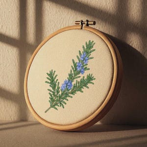 May include: A circular embroidery hoop with a beige fabric displaying a green and blue embroidered rosemary sprig. The wooden hoop is light brown, and the embroidery thread is a vibrant green and blue.