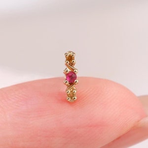 Puede incluir: Un pequeño pendiente de oro con una piedra preciosa de color rosa vibrante de corte redondo en el centro, flanqueada por dos esferas doradas más pequeñas. La pieza tiene un diseño vertical delgado y delicado con un acabado metálico pulido.