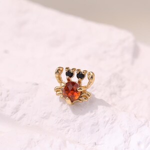 Puede incluir: Un pendiente de botón en tono dorado con forma de cangrejo, que presenta una piedra preciosa naranja facetada como cuerpo y dos pequeñas piedras redondas y oscuras como ojos. El diseño del cangrejo incluye pinzas y patas detalladas.