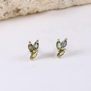 Puede incluir: Un par de pendientes de botón dorados, cada uno con tres hojas de piedras preciosas verdes. Los pendientes están colocados sobre un fondo blanco con una piedra beige texturizada en la parte superior de la imagen.