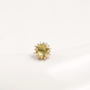 May include: A small, round, pale yellow gemstone stud earring featuring a textured, star-like metal setting with multiple pointed prongs radiating outward from the center, creating a sunburst effect on a smooth, light-colored surface.