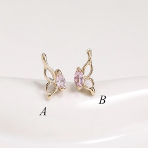 Puede incluir: Un par de pendientes dorados con forma de mariposa. Cada pendiente presenta una gema rosa de forma ovalada en el centro. Los pendientes se muestran sobre una superficie blanca, con las letras "A" y "B" al lado.