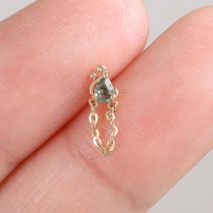 May include: A delicate gold-colored earring with a small, gray gemstone and a dangling chain. The earring is held between two fingers against a soft, neutral background. The jewelry piece is small and elegant.