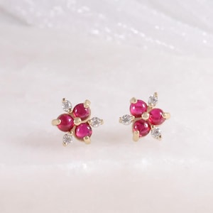 May include: A pair of gold-toned stud earrings, each featuring a flower design. The flower petals are a vibrant red, with small, clear accent stones. The earrings are displayed on a white surface.