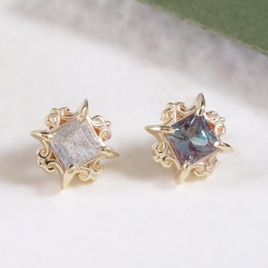 Puede incluir: Dos pendientes de botón dorados. Cada pendiente presenta una gema de corte cuadrado engastada en un diseño de estrella. Un pendiente tiene una gema gris, el otro una gema verde azulado. Los pendientes están sobre una superficie blanca.