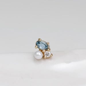 May include: A gold stud earring featuring an oval-cut blue gemstone, a small round white pearl, and a tiny round clear gemstone set in a cluster arrangement. The metal setting is yellow gold, and the stones are arranged in a compact, asymmetrical design.