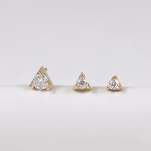 May include: Three gold-coloured triangle-shaped earrings with clear, sparkling stones. The earrings are arranged in a row, decreasing in size from left to right. The largest earring has a single, prominent stone, while the others have multiple smaller stones.