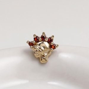 May include: A gold-toned skull-shaped stud earring featuring a crown-like arrangement of five small, round, deep red gemstones set along the top edge. The skull design includes detailed eye sockets and a nasal cavity, with a polished metallic finish.