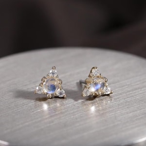 Könnte beinhalten: Ein Paar goldfarbene Ohrstecker. Jeder Ohrstecker hat ein dreieckiges Design mit einem zentralen, runden, irisierenden Stein, umgeben von kleineren, klaren Steinen. Die Ohrringe werden auf einer silberfarbenen, kreisförmigen Oberfläche präsentiert.