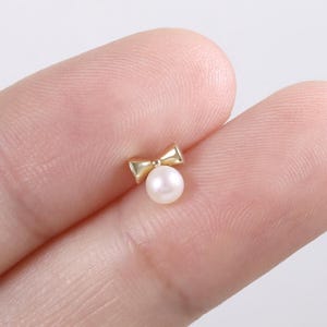 May include: A small, gold-colored bow-shaped earring with a single, round, white pearl. The earring is shown against a light background.