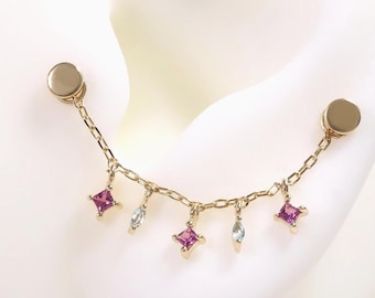 May include: Gold-toned ear jewelry featuring a chain connecting two round studs. The chain is adorned with small, dangling charms, including pink square-shaped gems and clear, teardrop-shaped gems. The jewelry is displayed on a white surface.