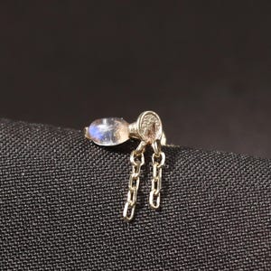 May include: A small gold-toned earring featuring a single oval-shaped iridescent moonstone cabochon set in metal. Two delicate, short gold-toned metal chains hang vertically from the base of the stone setting, creating a dangling effect.